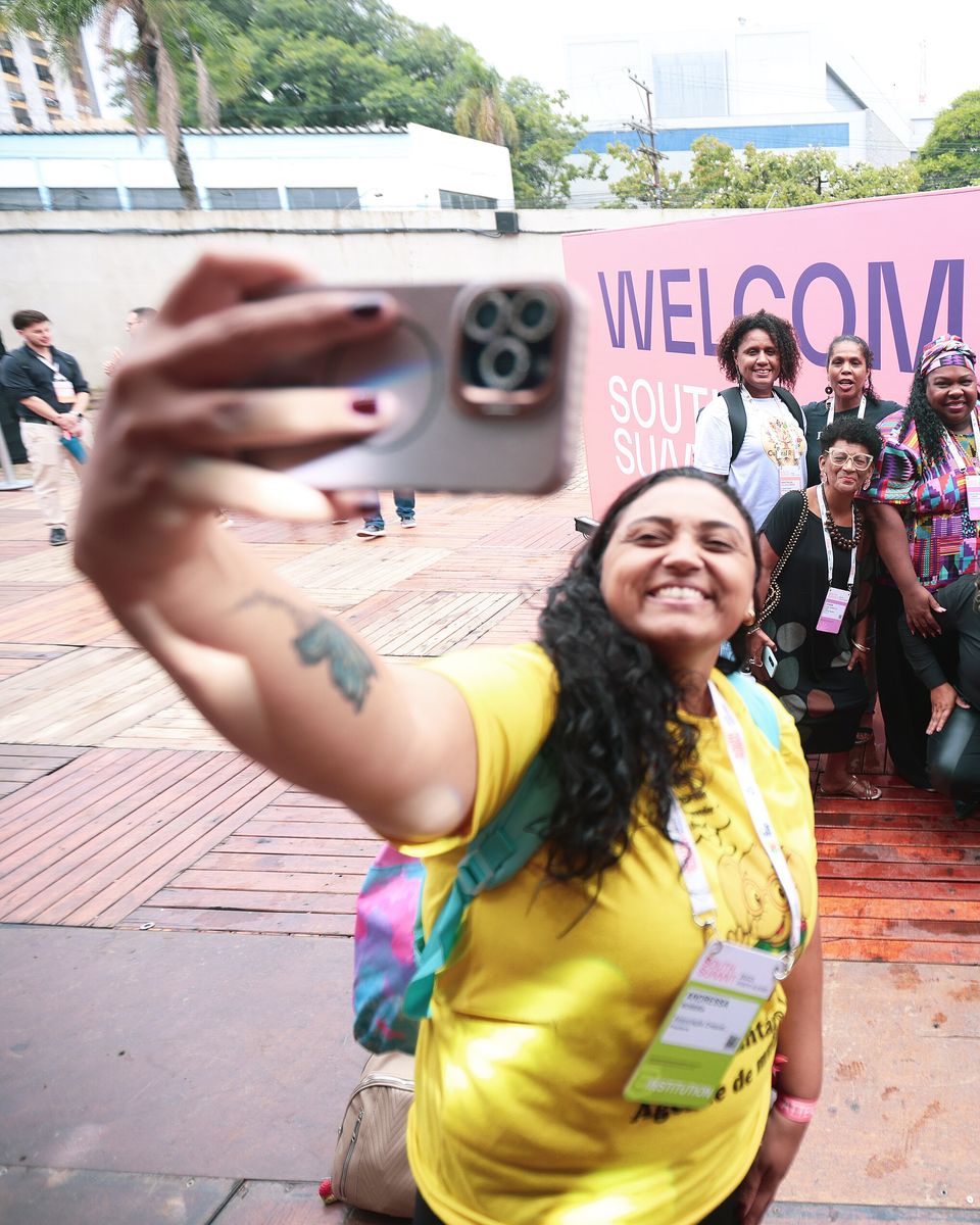 Participante de camiseta amarela e crachá tira uma selfie com o celular em frente a um painel rosa na entrada do South Summit Brazil, em Porto Alegre, com um grupo de mulheres sorrindo atrás dela.