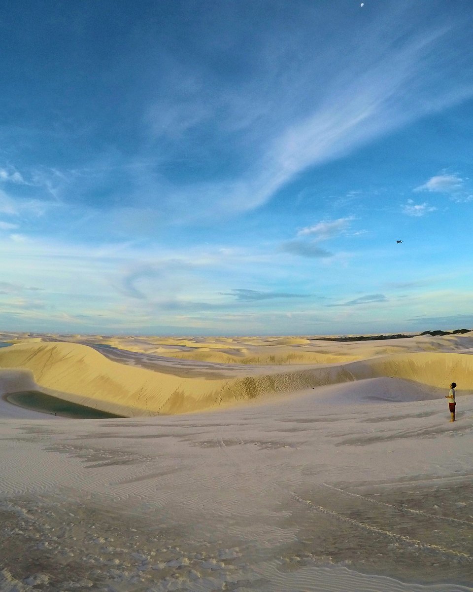 Visitante sozinho em pé sobre uma duna dos Lençóis Maranhenses no fim da tarde, com uma lagoa à esquerda, campo de dunas até o horizonte e um drone pequeno no céu azul.