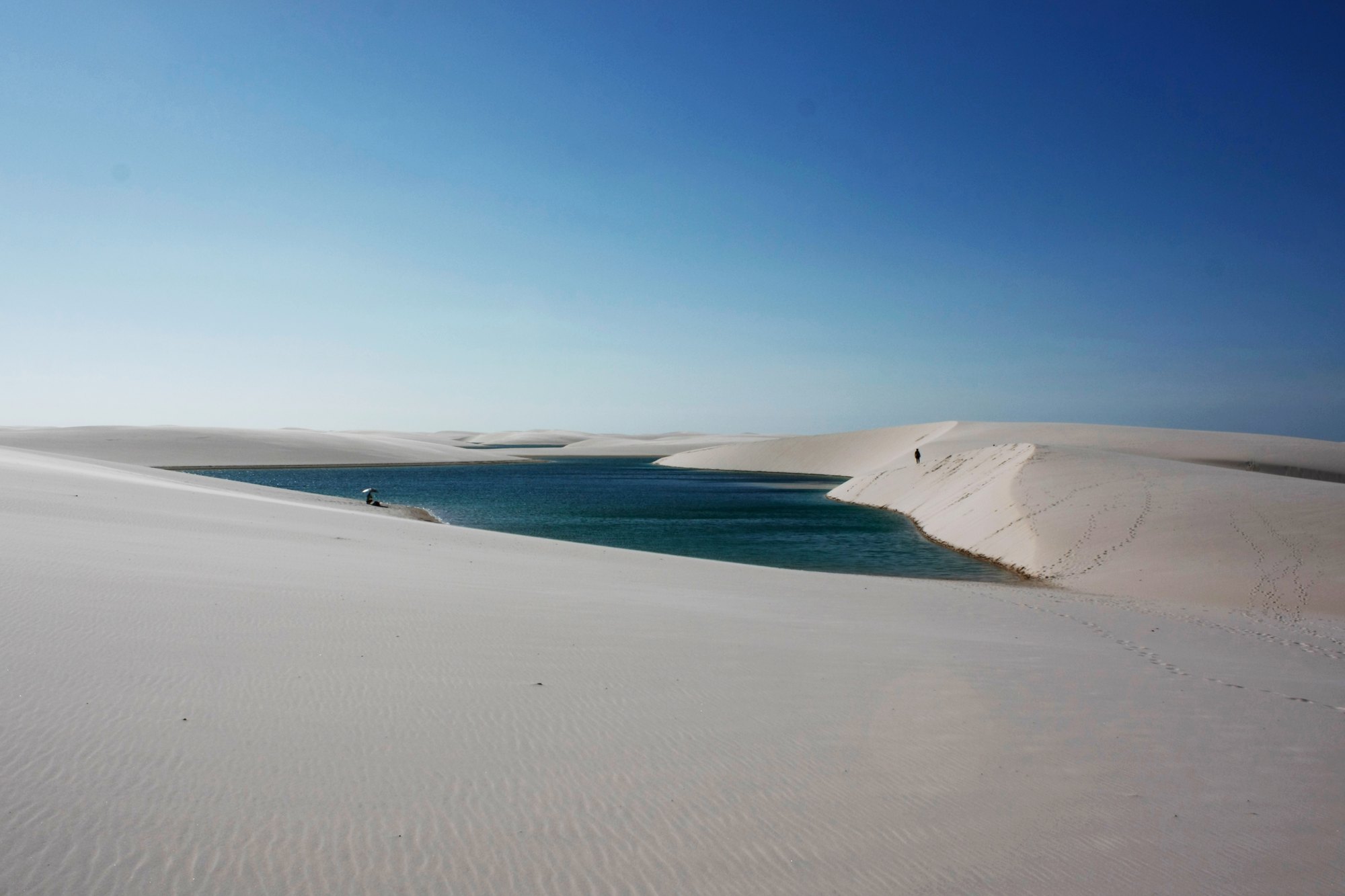 Lagoa comprida de água verde-azulada encaixada entre dunas de areia branca no Parque Nacional dos Lençóis Maranhenses, com duas pessoas ao longe na crista da duna e rastros de pegadas na areia.