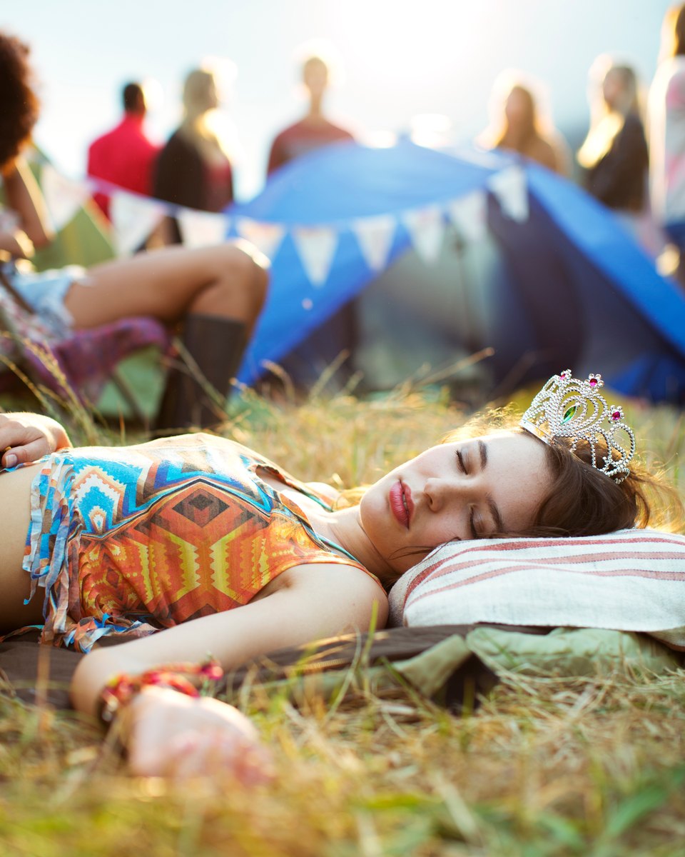 Jovem de tiara e top estampado dormindo na grama, com a cabeça num travesseiro, diante de barracas de camping e bandeirolas num festival de música.