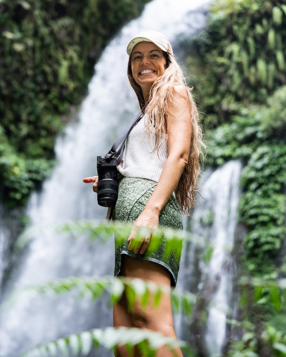 Mulher de boné e cabelo molhado sorrindo com uma câmera pendurada no ombro, diante de uma cachoeira no meio da mata.