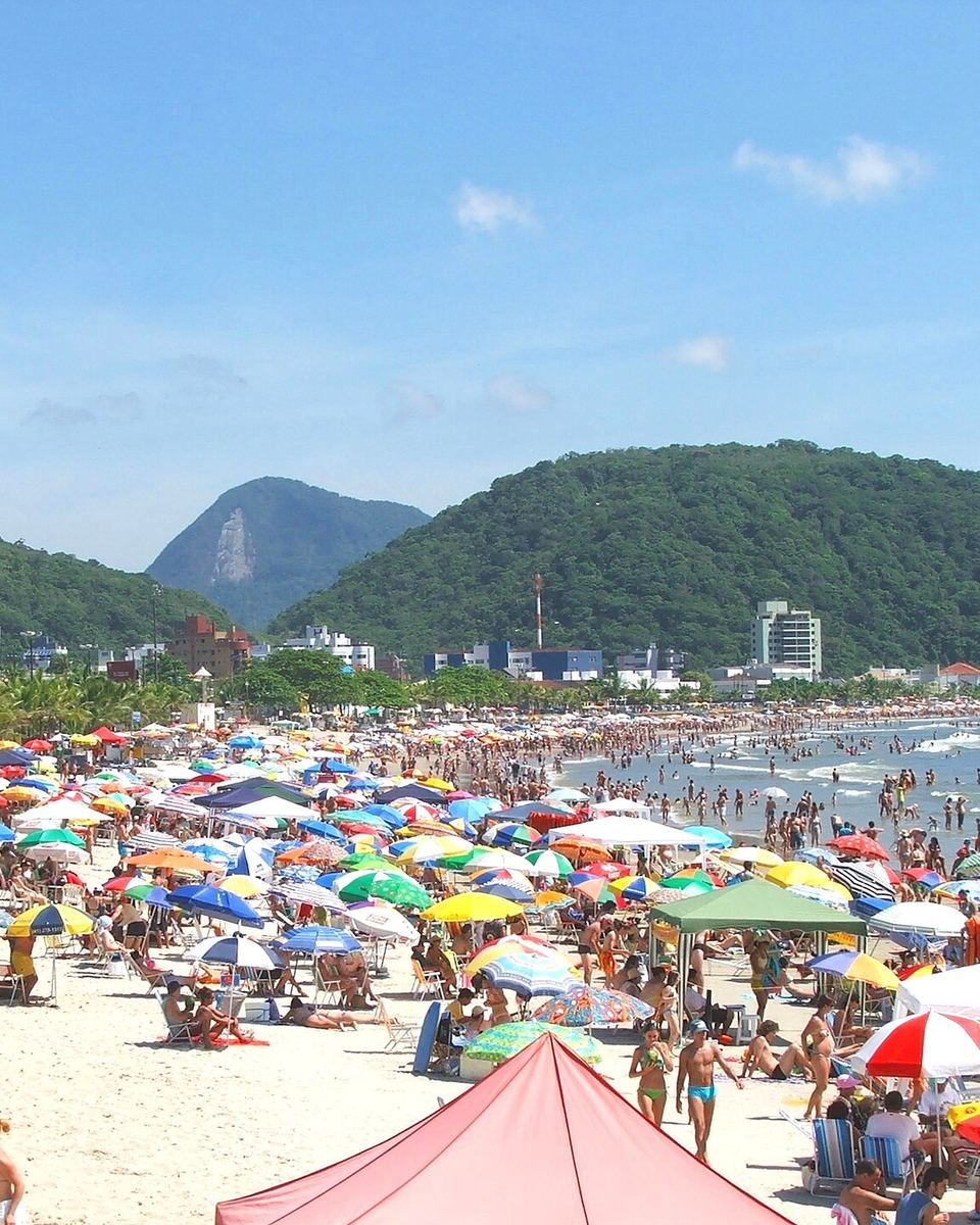 Praia Central de Guaratuba lotada num dia de sol, com guarda-sóis coloridos na areia, banhistas no mar e morros cobertos de mata ao fundo.