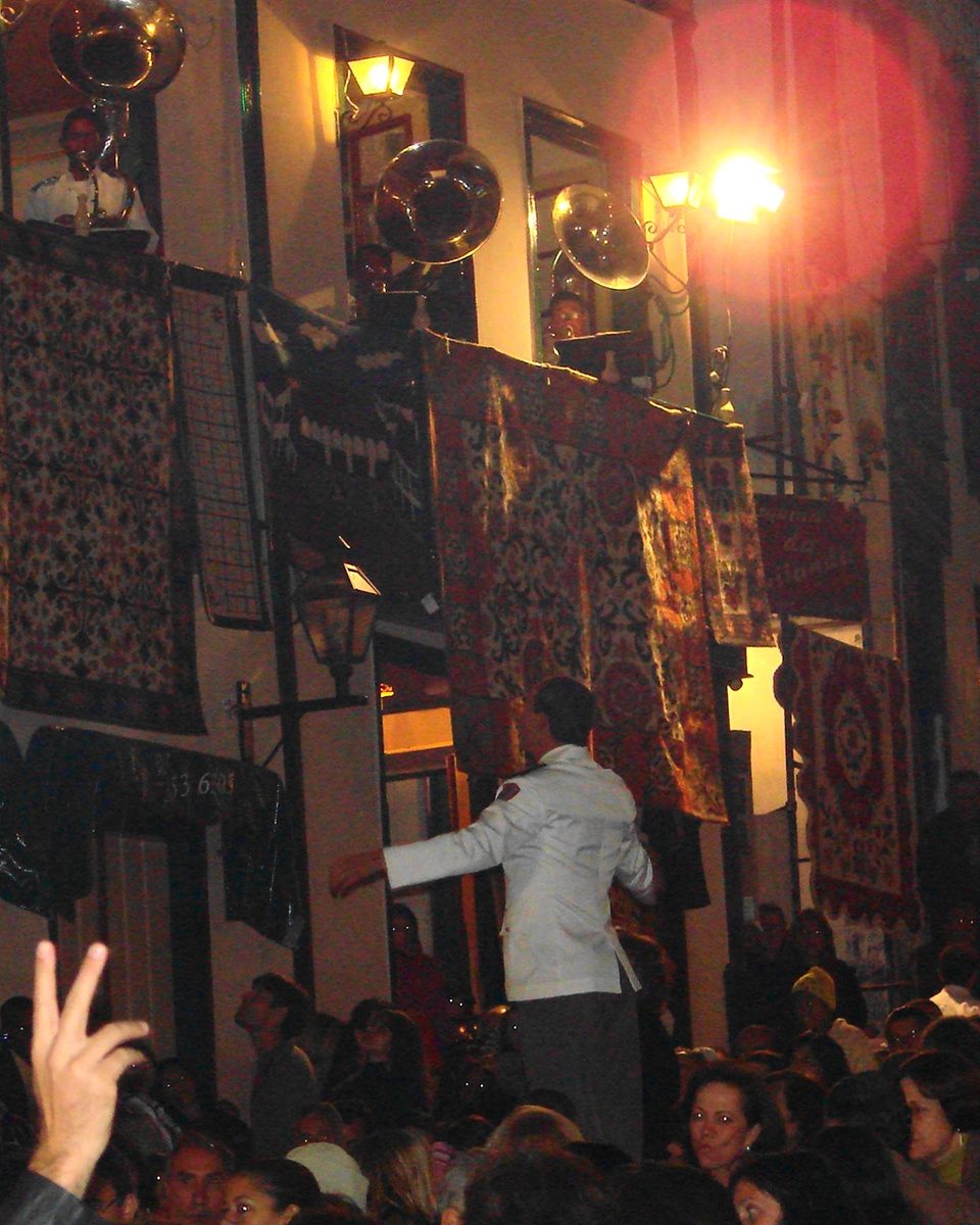 Maestro de paletó branco regendo no meio do público, à noite, na Rua da Quitanda em Diamantina, enquanto músicos com tuba e trompete tocam na sacada de um casarão coberta de tapeçarias.