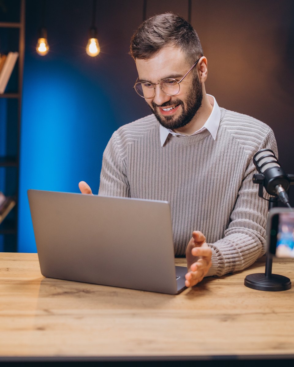 Homem de óculos e suéter fala sorrindo diante de um notebook numa mesa de madeira, com um microfone ao lado e um celular num tripé gravando a cena.