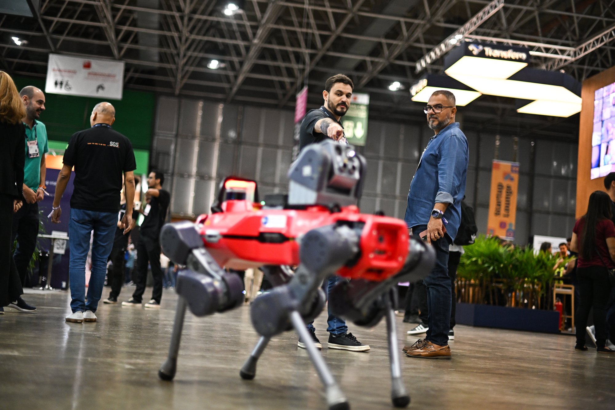 Participantes circulam entre os estandes de startups no pavilhão do Riocentro, no Rio de Janeiro, enquanto um robô quadrúpede vermelho anda em primeiro plano.