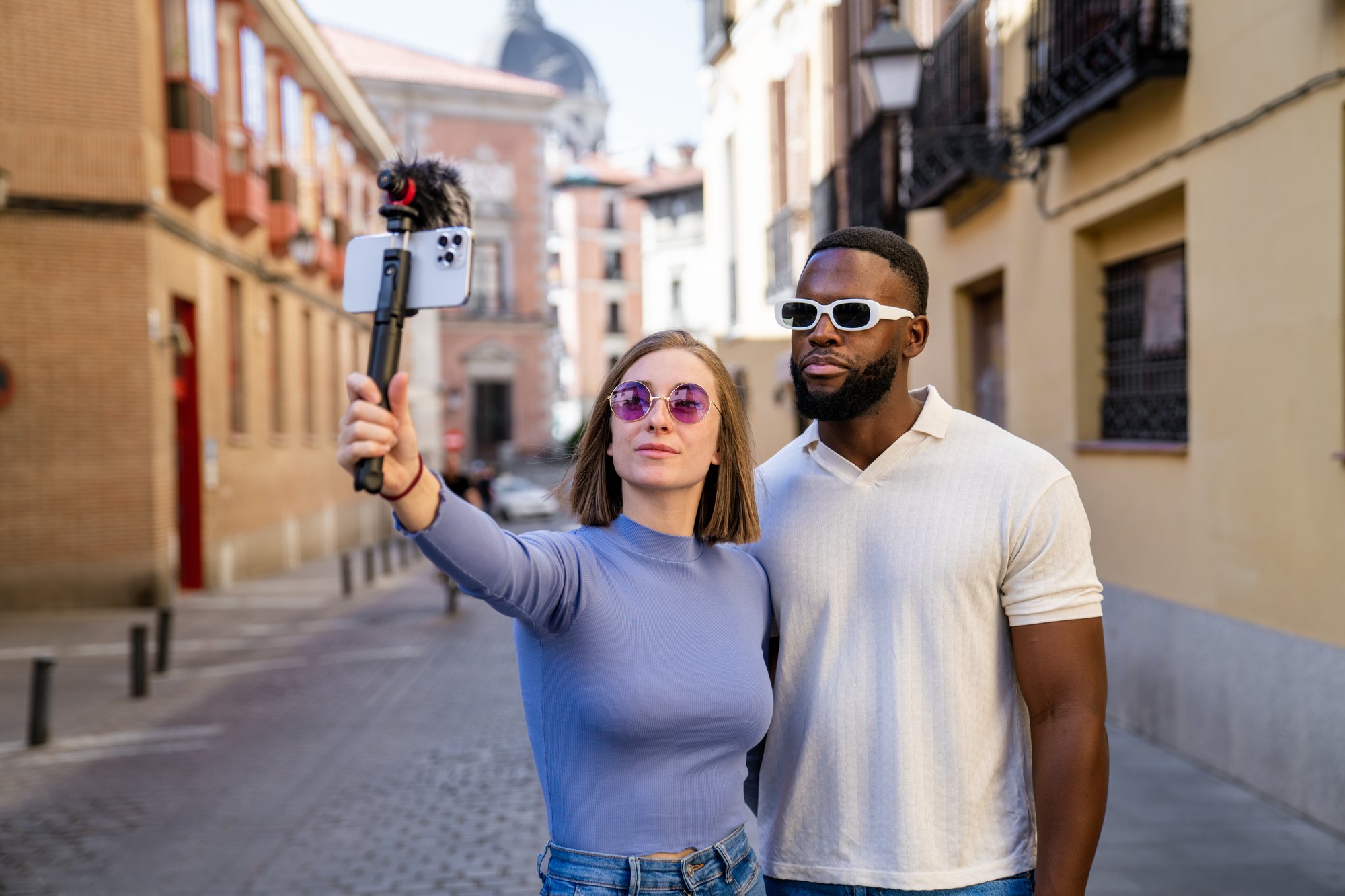 Casal gravando vídeo de viagem com o celular num estabilizador, com microfone, numa rua de casarões antigos.