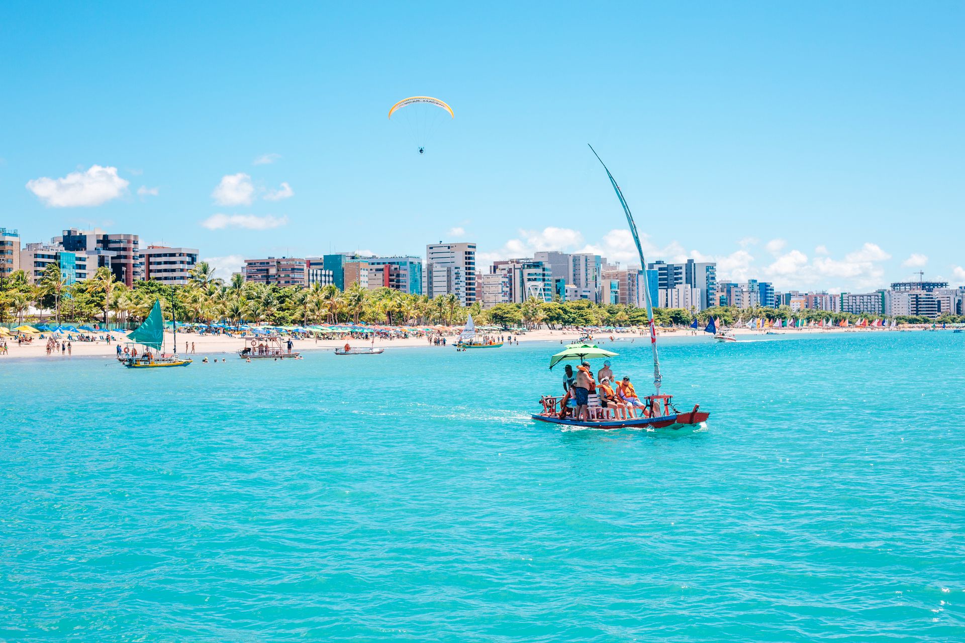 Barco navegando sobre as águas turquesa da orla de Maceió, com os prédios da cidade ao fundo