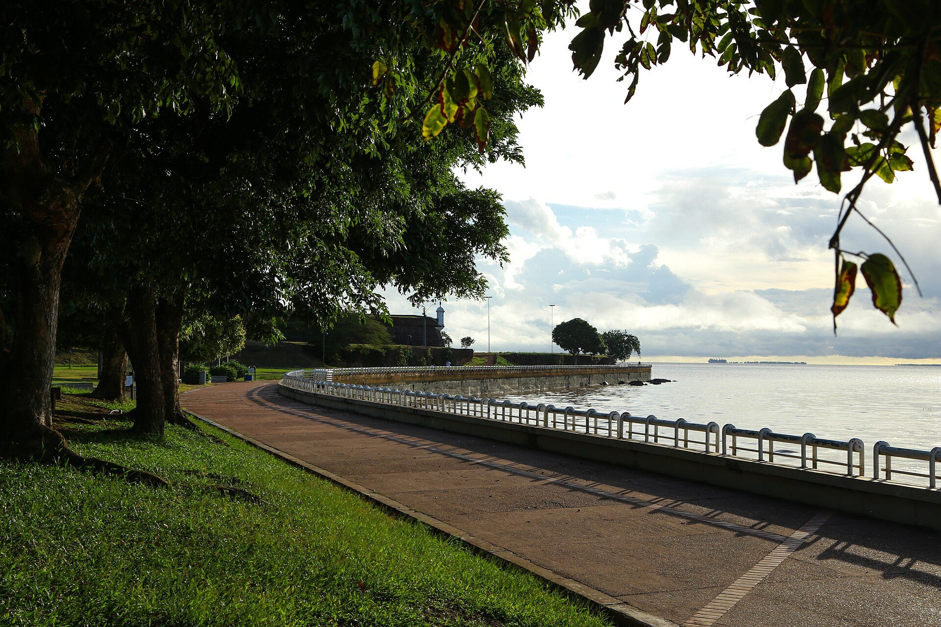 Orla de Macapá ao amanhecer, com o calçadão sob árvores, o muro de pedra sobre o rio Amazonas e a silhueta da Fortaleza de São José ao fundo