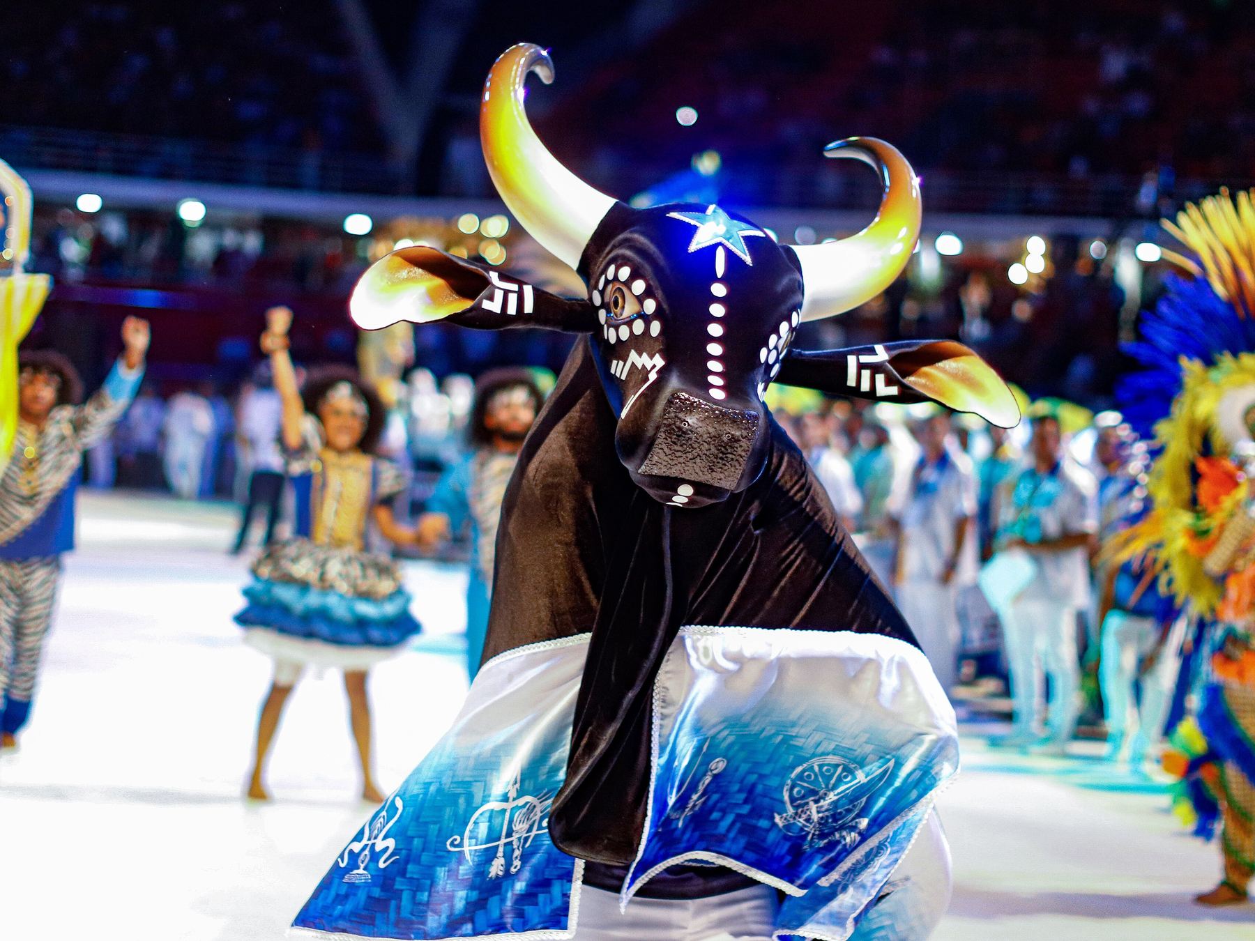 Apresentação noturna no bumbódromo de Parintins, com a figura do boi de chifres brancos dançando no centro da arena e brincantes fantasiados ao redor