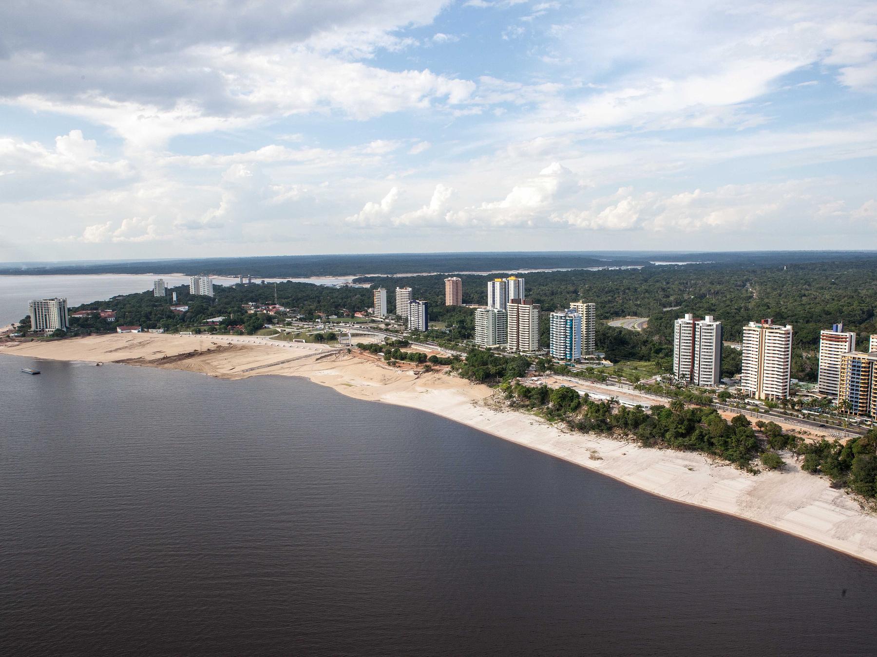 Vista aérea da Praia da Ponta Negra em Manaus, com a faixa de areia clara acompanhando a margem do rio Negro e os prédios do bairro entre a mata