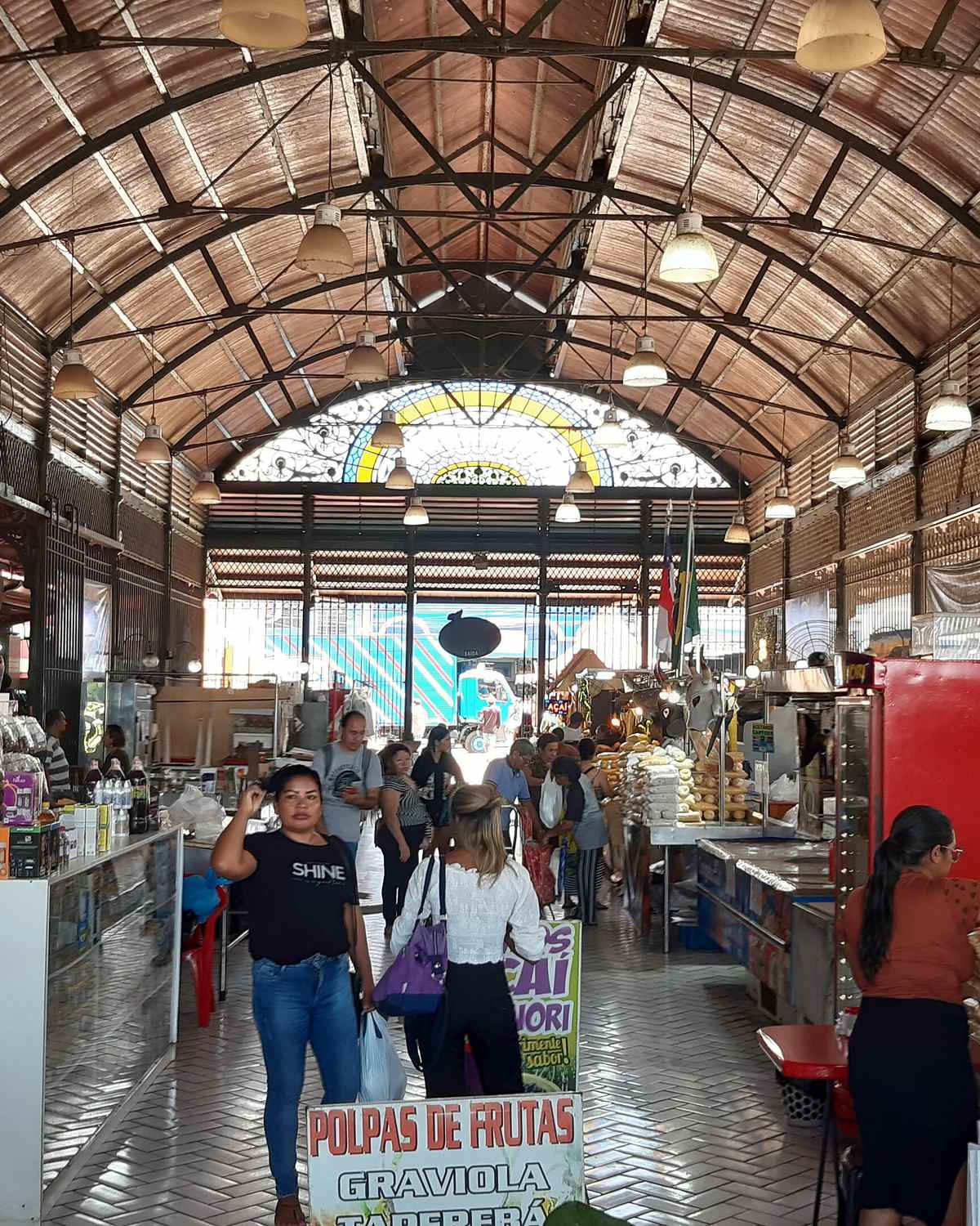 Interior do Mercado Municipal Adolpho Lisboa, em Manaus, com a estrutura de ferro e o vitral ao fundo, bancas de temperos e frutas e pessoas circulando entre os corredores.