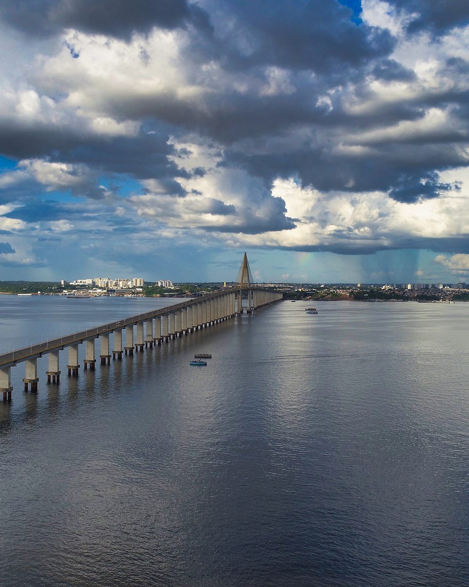 Vista aérea da Ponte Rio Negro ligando Manaus a Iranduba, com a pista curvando sobre os pilares, a torre estaiada ao fundo e nuvens altas sobre a cidade.