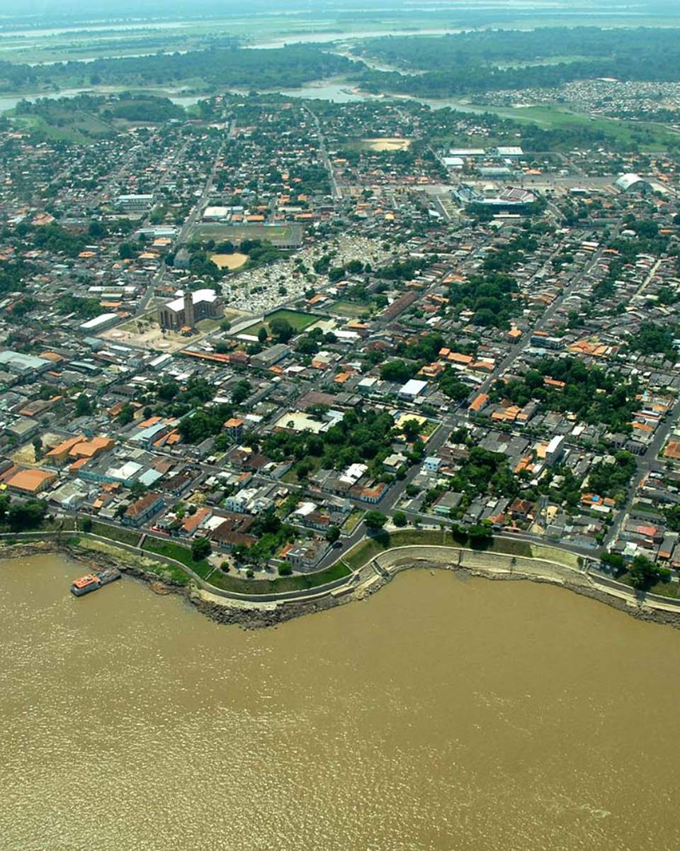 Vista aérea da cidade de Parintins às margens do rio Amazonas, com as ruas e casas do centro, a orla de pedras e barcos atracados.