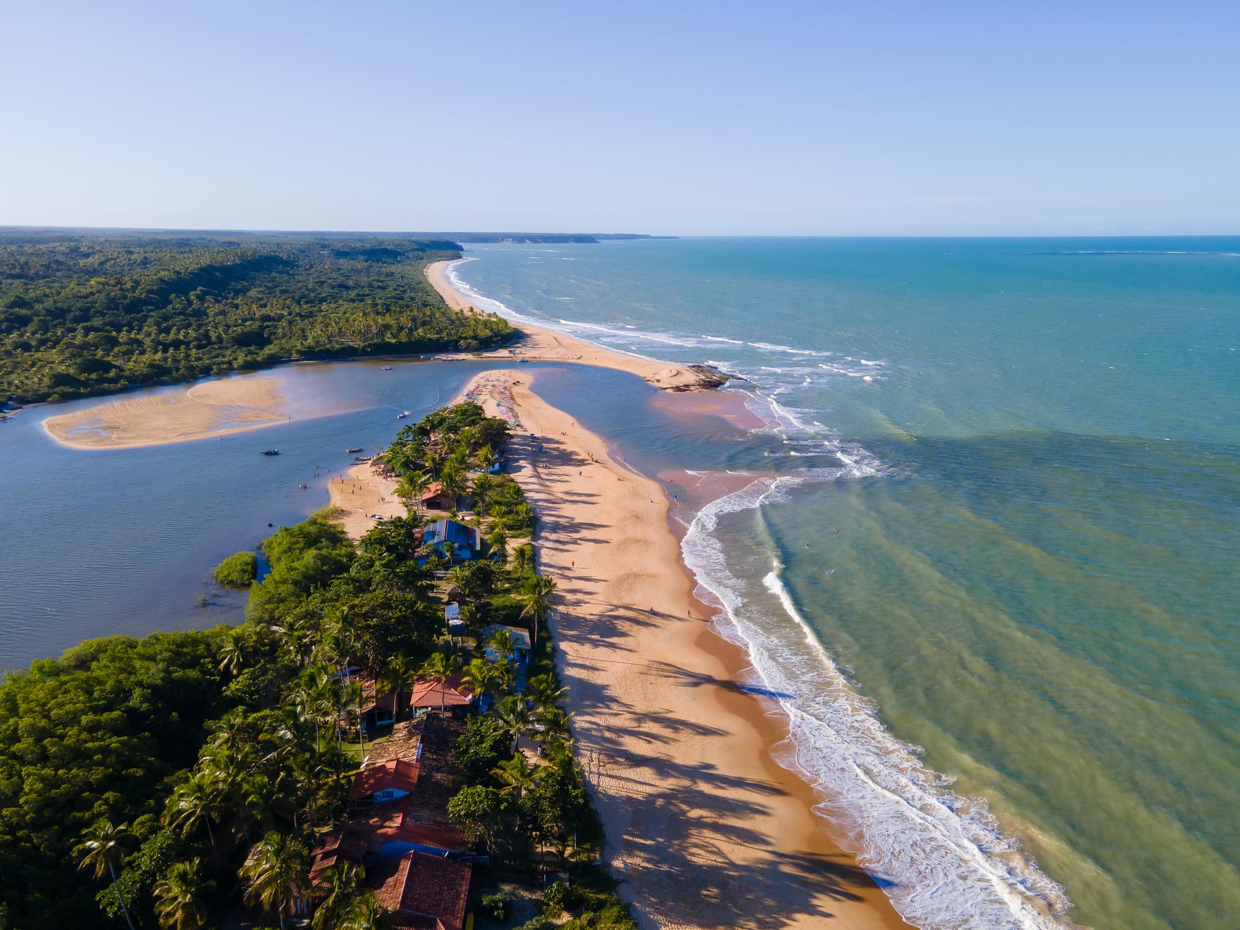 Vista aérea de Caraíva, com a vila entre o rio e o mar, a língua de areia na foz e a mata atlântica ao redor.