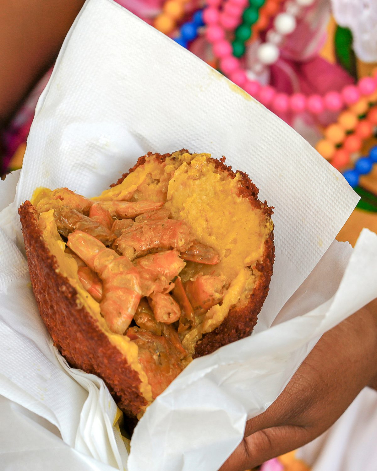 Acarajé aberto com vatapá e camarão, servido num papel branco, nas mãos de uma baiana de pulseira dourada e colares de contas coloridas.