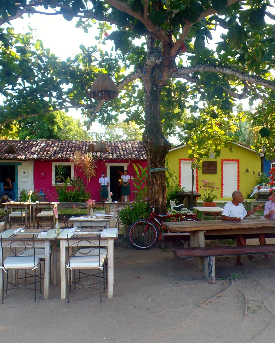 Casas coloridas com mesas e cadeiras ao ar livre sob árvores grandes no Quadrado de Trancoso, com pessoas conversando à sombra.