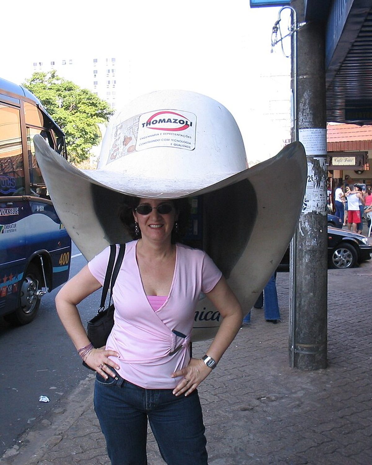 Turista de camiseta rosa e óculos escuros posando embaixo de um orelhão em forma de chapéu de peão gigante, numa calçada do Centro de Barretos, com um ônibus de turismo estacionado ao lado.