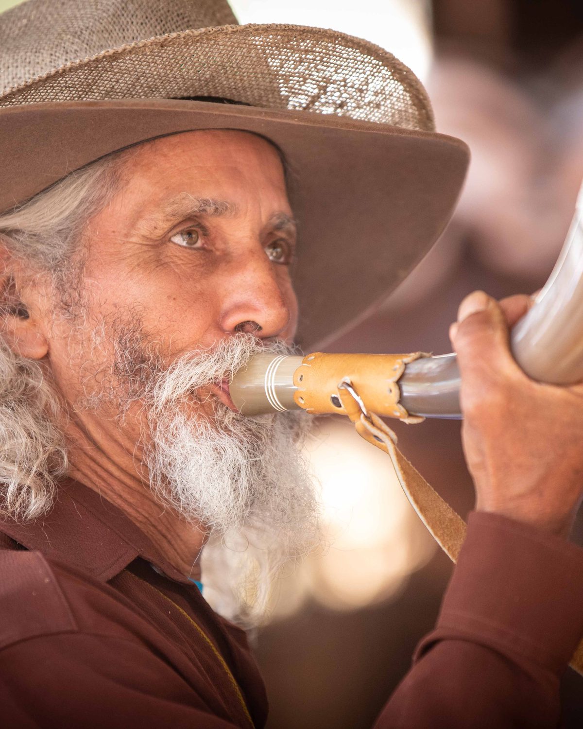 Peão de cabelos compridos e barba brancos, chapéu marrom de aba larga e camisa marrom, tocando um berrante de chifre com bocal de couro, na Queima do Alho em Barretos.