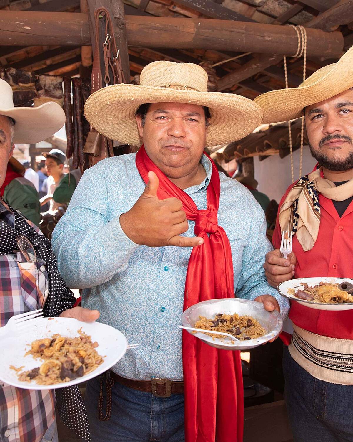Três homens de chapéu de palha, camisa xadrez e lenço no pescoço sorrindo com pratos de comida na mão, num rancho de madeira na Queima do Alho, em Barretos.
