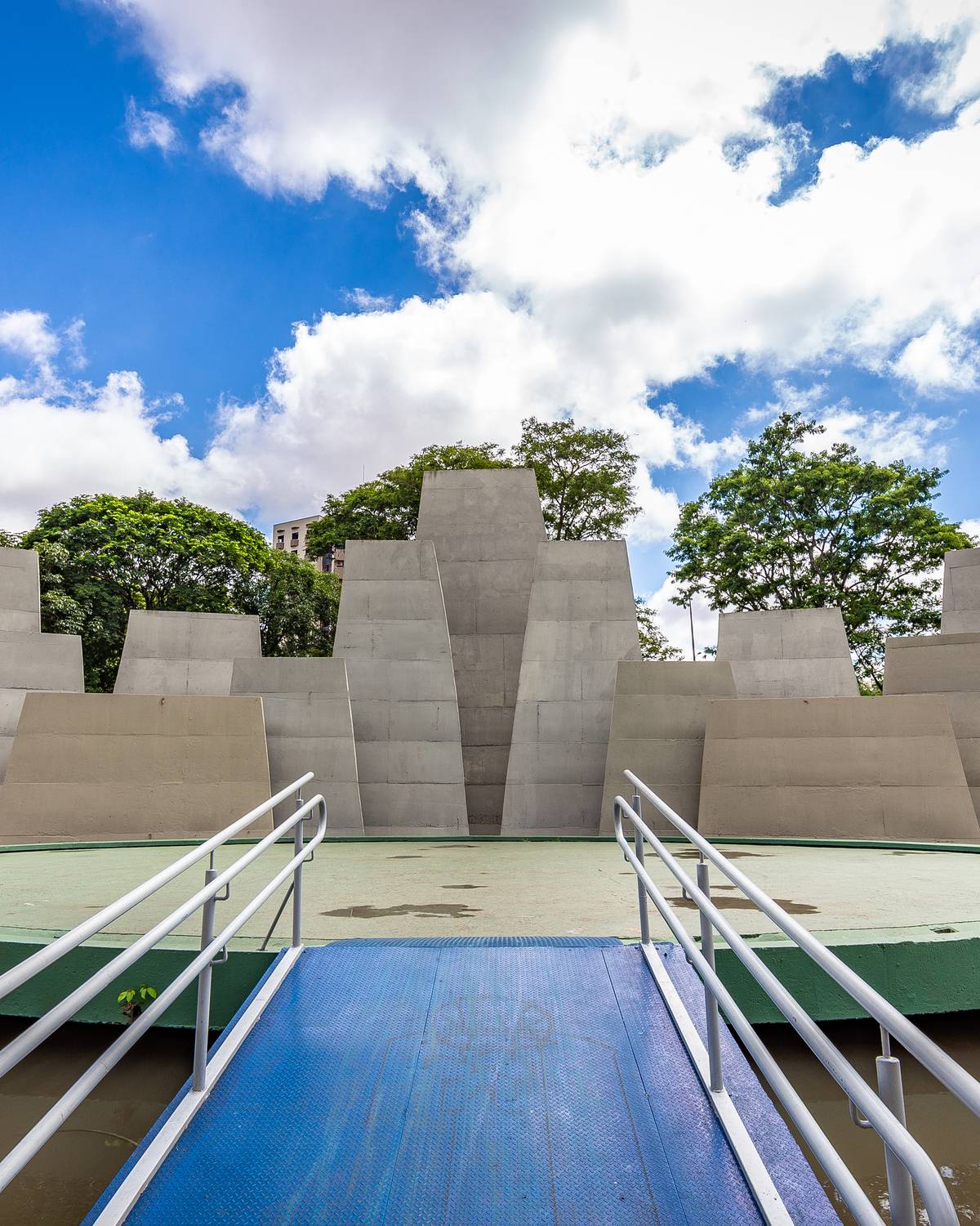 Anfiteatro ao ar livre do Parque Vitória Régia, em Bauru, com a concha de concreto sobre o lago, passarela de acesso e árvores ao redor.