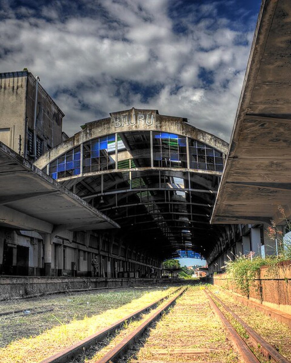 Plataforma da Estação Ferroviária de Bauru, com a cobertura de concreto, os trilhos tomados pelo mato e o prédio da estação ao fundo, sob céu azul com nuvens.