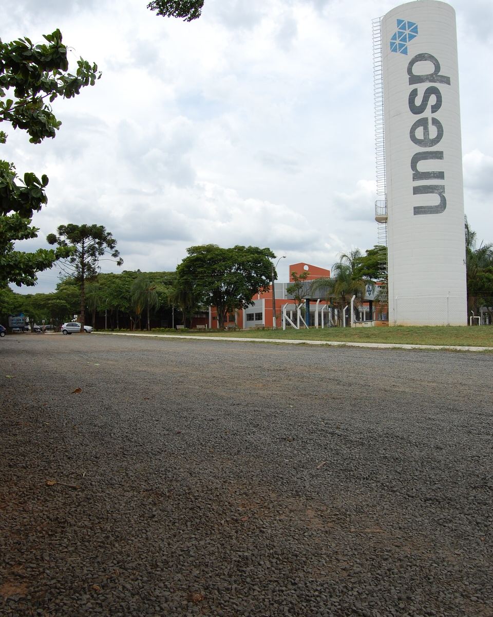 Entrada do câmpus da Unesp em Bauru, com a torre branca de letreiro vertical da universidade à direita, árvores e a avenida de acesso vazia.