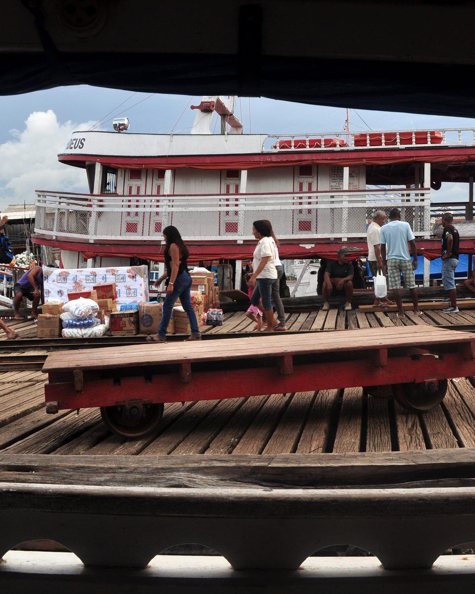 Barco de linha branco e vermelho atracado no cais da Cidade Velha, em Belém, com pessoas carregando caixas e sacos pela prancha de madeira.