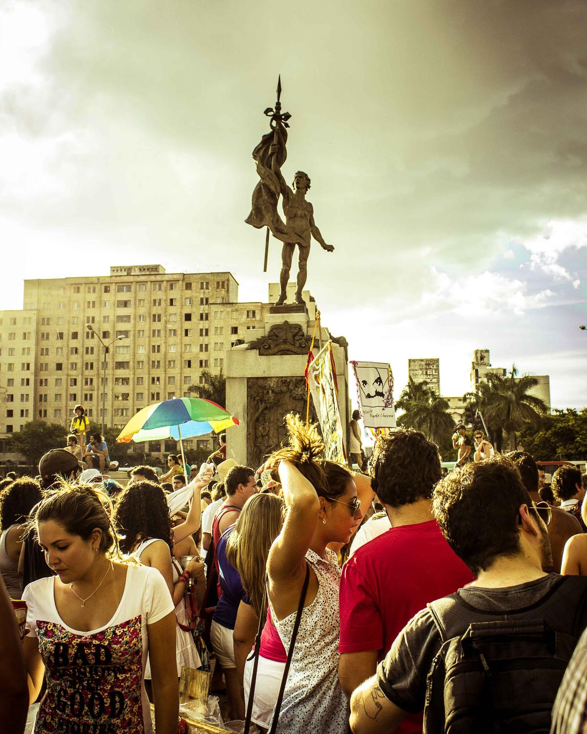 Multidão na Praça da Estação, no Centro de Belo Horizonte, durante a Praia da Estação, com um guarda-sol colorido e o monumento da praça contra o céu do fim de tarde.