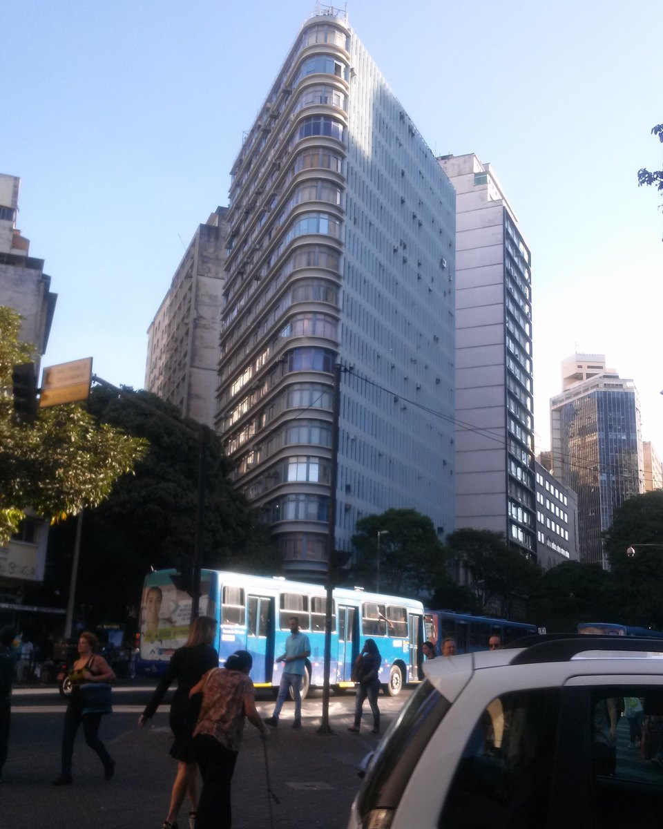 Pedestres atravessando a rua na Praça Sete, no Hipercentro de Belo Horizonte, com um ônibus azul passando diante de prédios altos no fim da tarde.