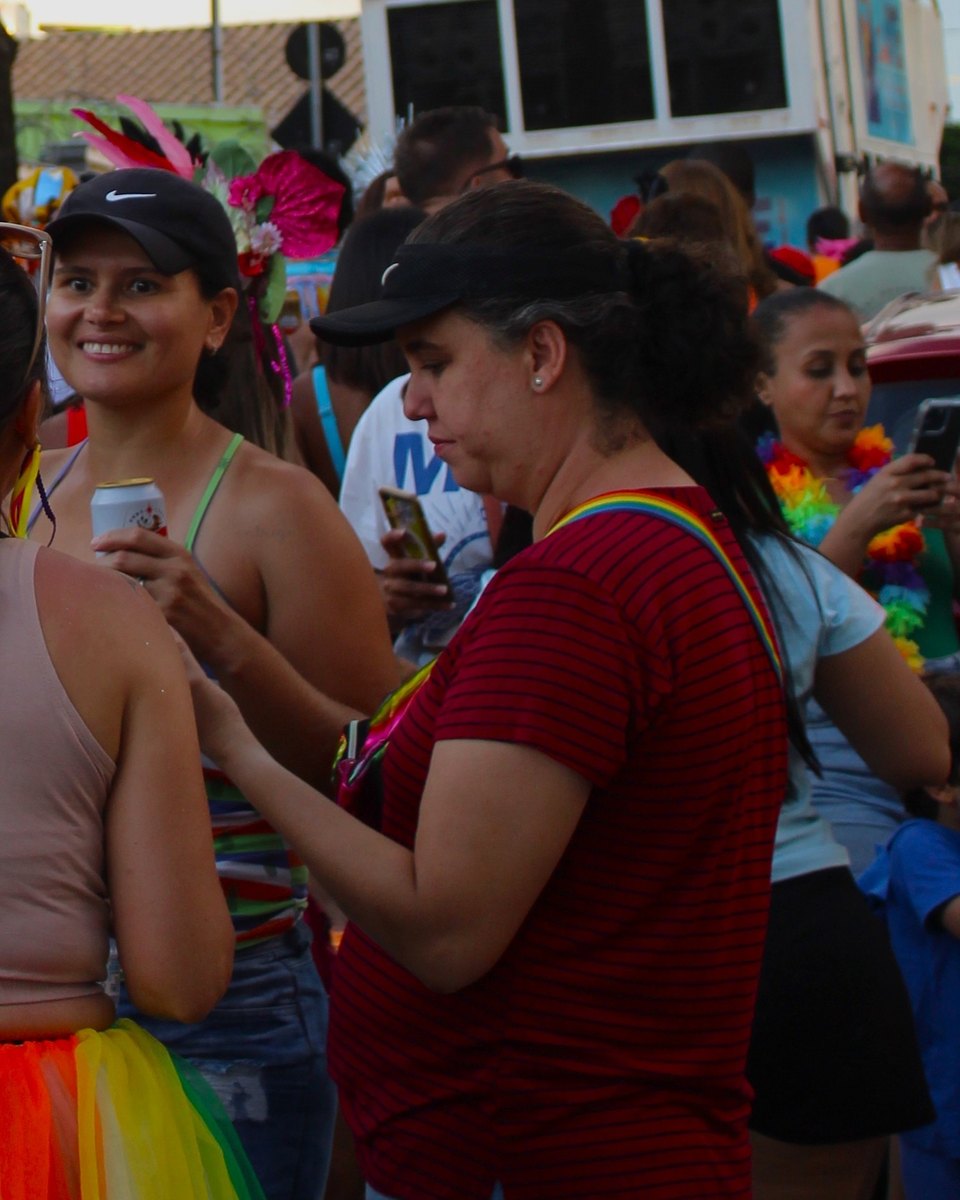 Foliãs de boné e colar colorido no bloco da Graça, no Carnaval de Belo Horizonte, uma sorrindo com uma lata na mão e outra olhando o celular, com o trio elétrico ao fundo.