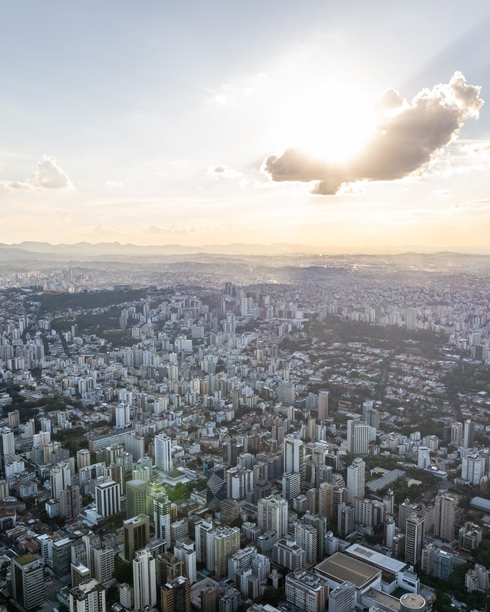 Vista aérea de Belo Horizonte no fim da tarde, com prédios e bairros se espalhando até as serras no horizonte e o sol atrás de uma nuvem.