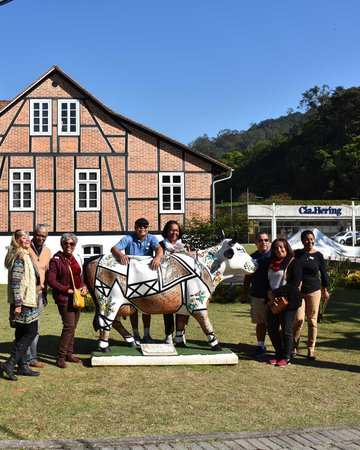 Grupo de visitantes posando ao lado de uma escultura de vaca pintada no gramado em frente a uma casa enxaimel de tijolos, no Museu Hering, em Blumenau, num dia de céu azul.