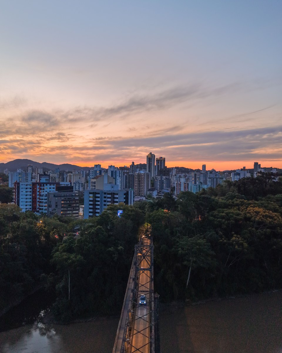 Vista aérea da Ponte de Ferro sobre o rio Itajaí-Açu, em Blumenau, ao pôr do sol, com a mata nas margens e os prédios da cidade contra o céu alaranjado.