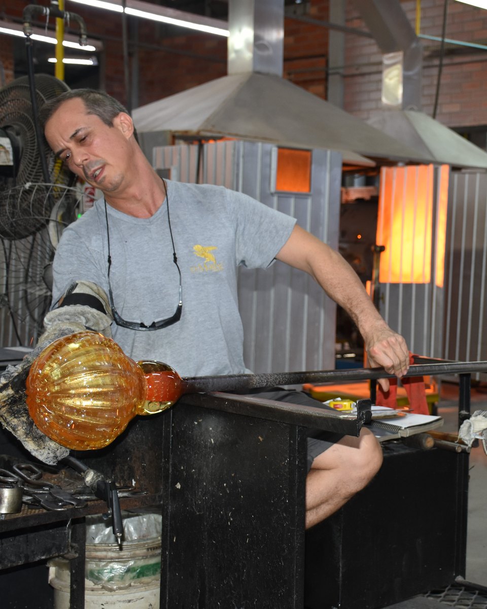 Artesão soprando uma peça de cristal alaranjada na ponta de um tubo de metal, ao lado do forno, na fábrica do GlasPark Museu do Cristal, em Blumenau.