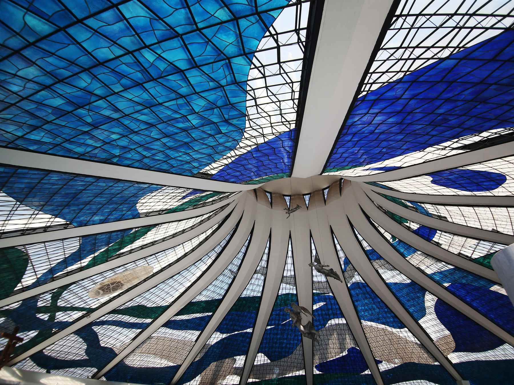 Interior da Catedral Metropolitana de Brasília visto de baixo, com as colunas curvas de concreto subindo até o topo e os vitrais azuis, brancos e verdes preenchendo todo o teto.