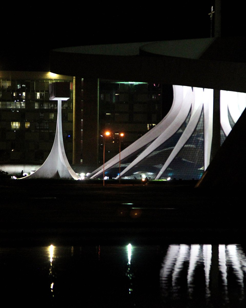 Catedral Metropolitana de Brasília iluminada à noite, com as colunas curvas de concreto e o campanário acesos, prédios ao fundo e as luzes refletidas na água em primeiro plano.