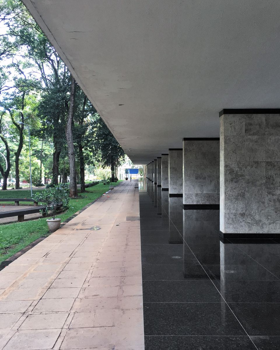 Passagem sob os pilotis de um bloco residencial de superquadra em Brasília, com as colunas de concreto, o piso de ladrilho e as árvores do gramado aparecendo do outro lado.
