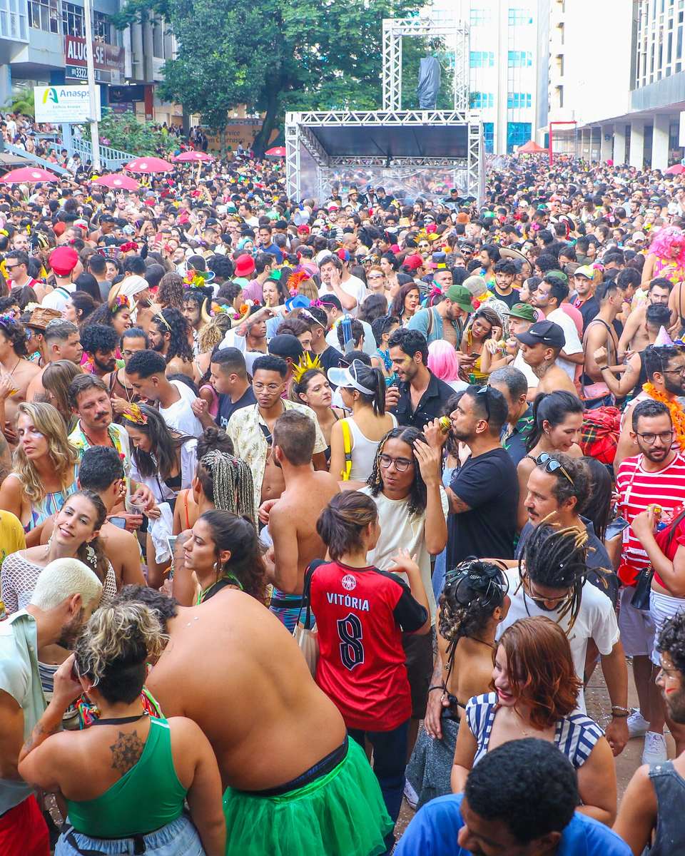 Rua do Setor Comercial Sul, em Brasília, tomada por uma multidão de gente fantasiada entre os prédios de escritório, com palco de treliça metálica ao fundo.