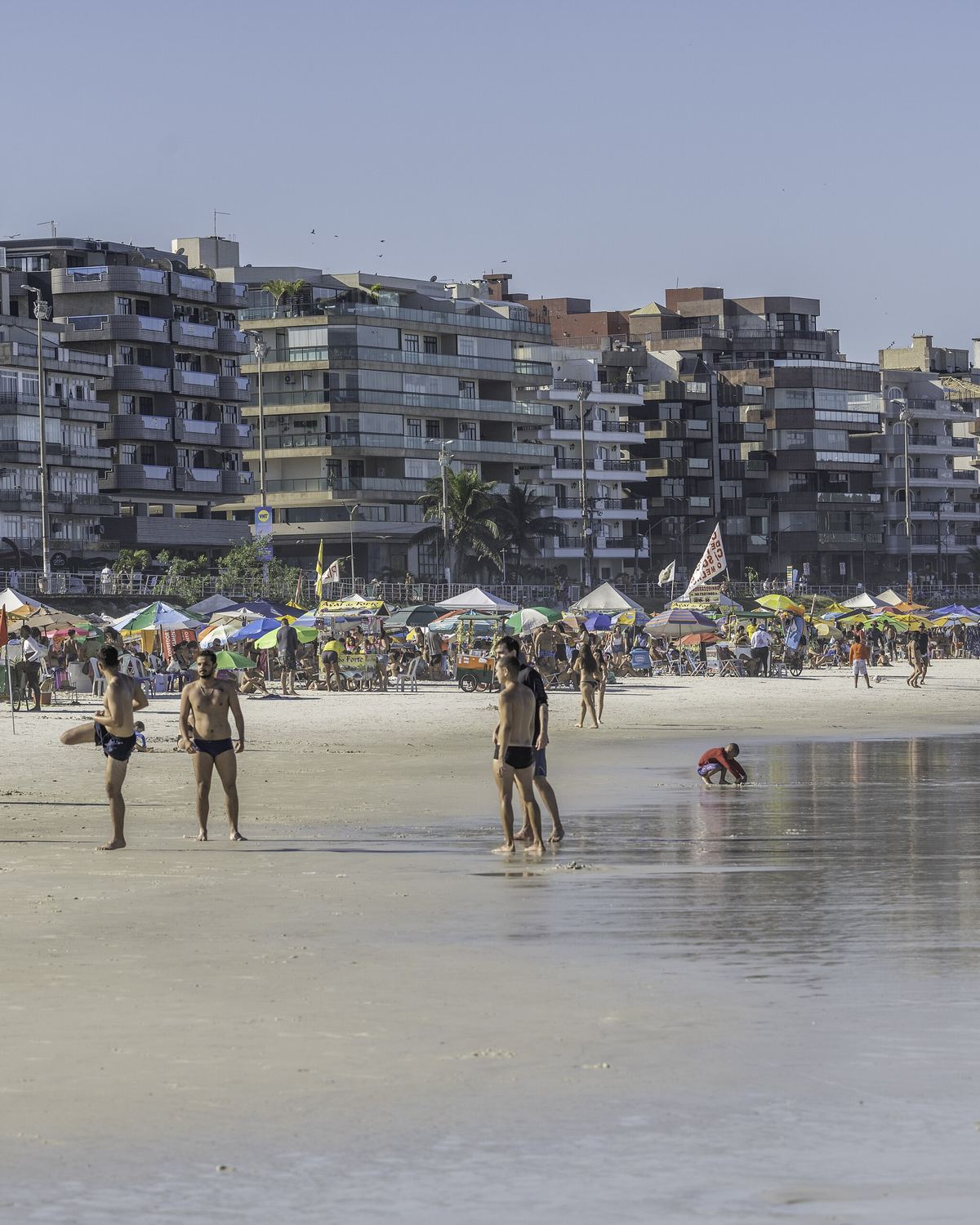 Banhistas caminhando na areia molhada da Praia do Forte, em Cabo Frio, com guarda-sóis e a fileira de prédios da orla ao fundo.