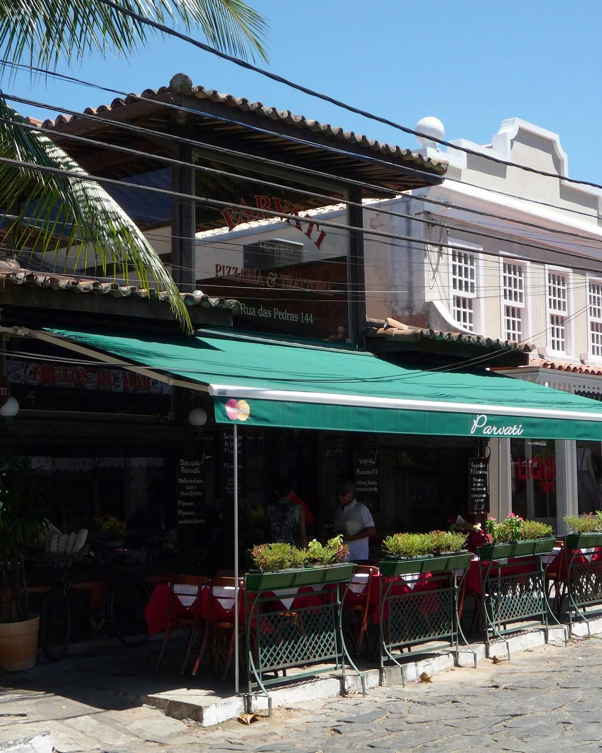 Restaurante com toldo verde, mesas de toalha vermelha e floreiras na calçada de pedra da Rua das Pedras, em Búzios, com um sobrado branco ao lado e um homem passando na rua num dia de sol.