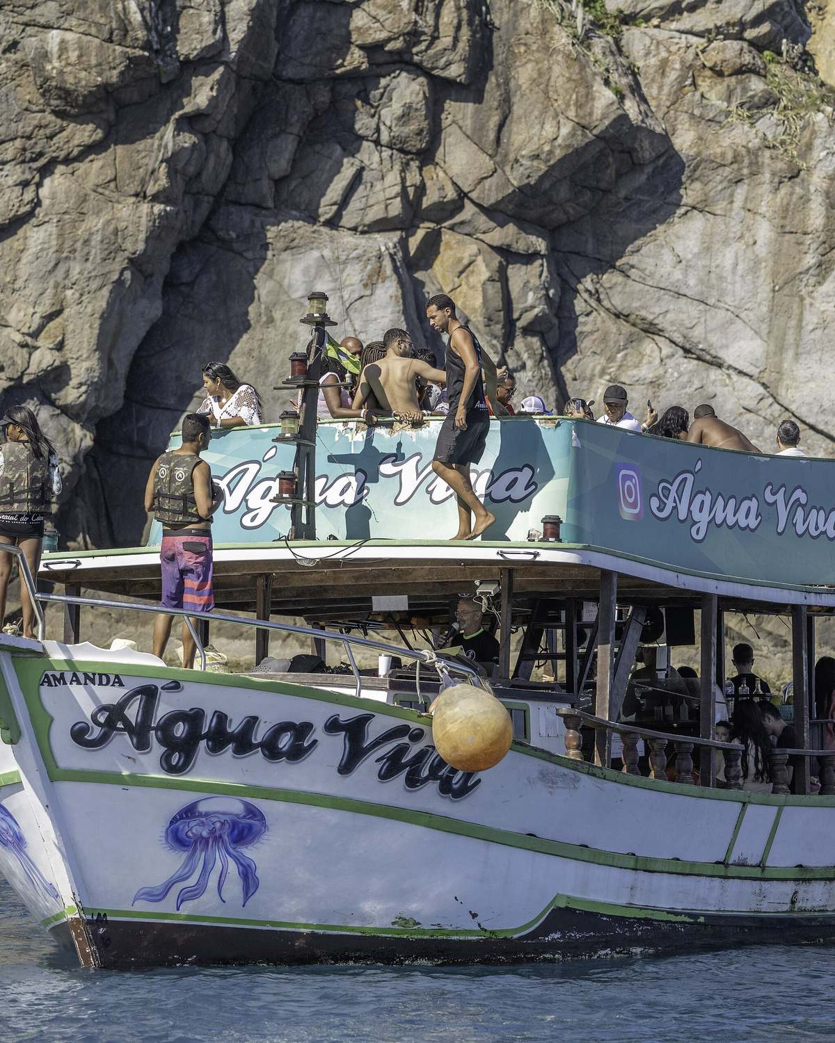 Barco de passeio cheio de turistas ancorado junto a um paredão de pedra, em Arraial do Cabo, com gente no deck de cima pronta para pular na água.