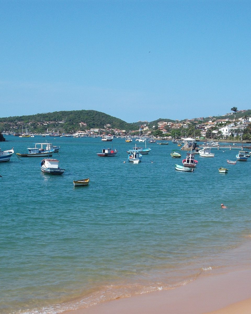 Barcos de pesca e de passeio ancorados na água azul de uma enseada de Búzios, com banhistas na parte rasa, a areia dourada à direita e morros com casas ao fundo.