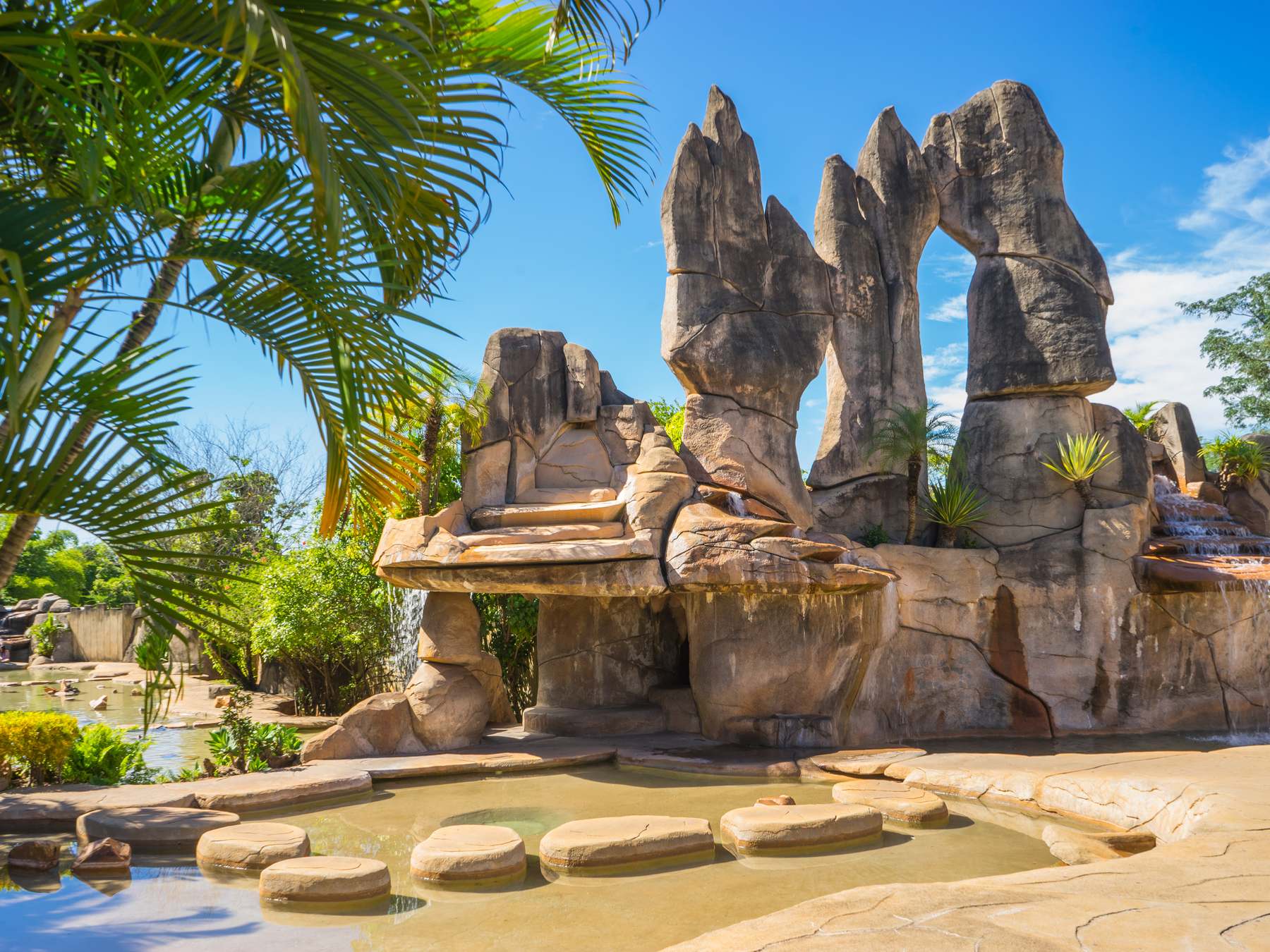 Monumento das Águas, em Caldas Novas, com grandes esculturas de pedra em forma de rochedos e um arco, cascatas e um espelho d'água com pedras de passagem, entre palmeiras e céu azul.
