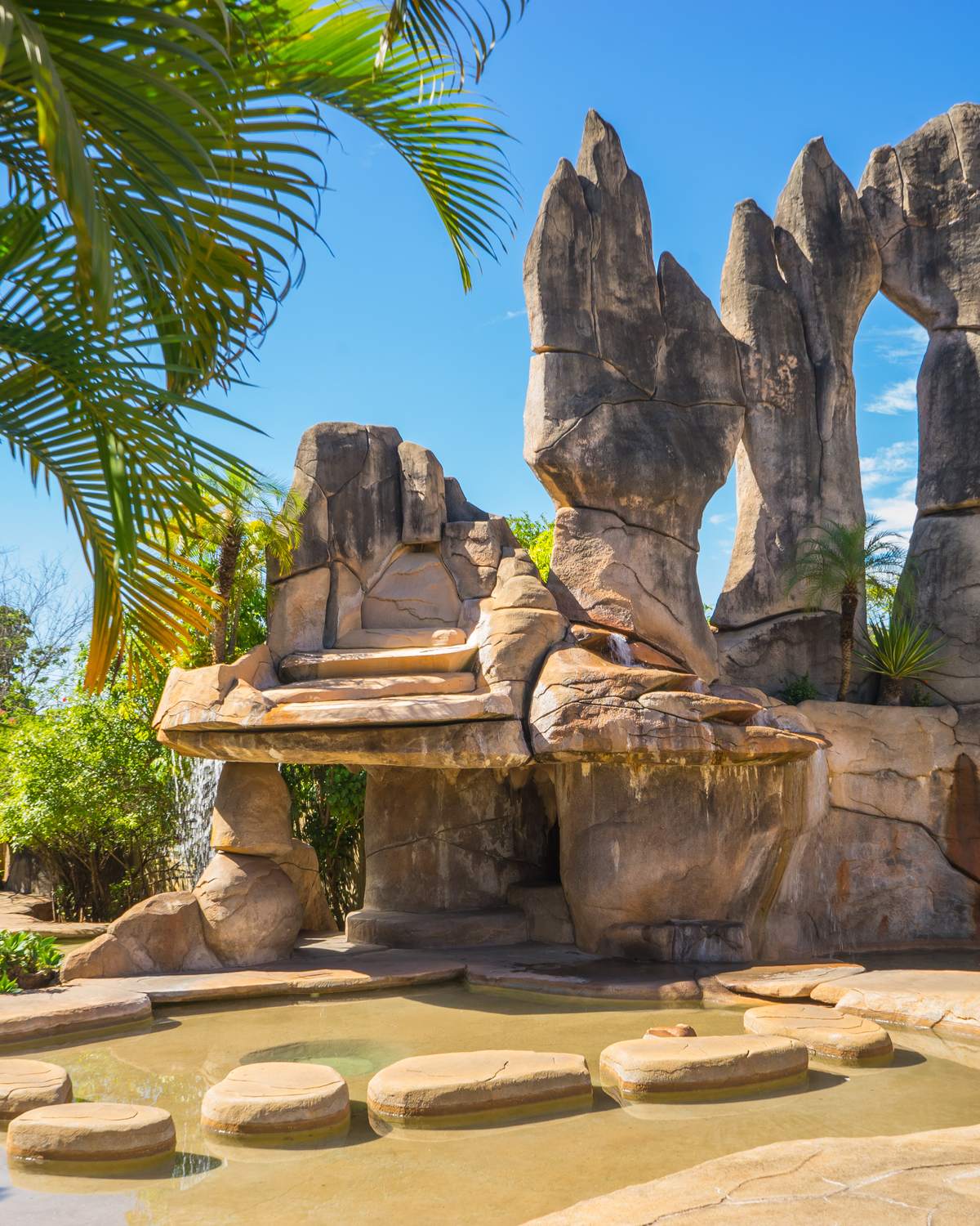 Monumento das Águas, em Caldas Novas, com grandes esculturas de pedra em forma de rochedos e um arco, cascatas e um espelho d'água com pedras de passagem, entre palmeiras e céu azul.
