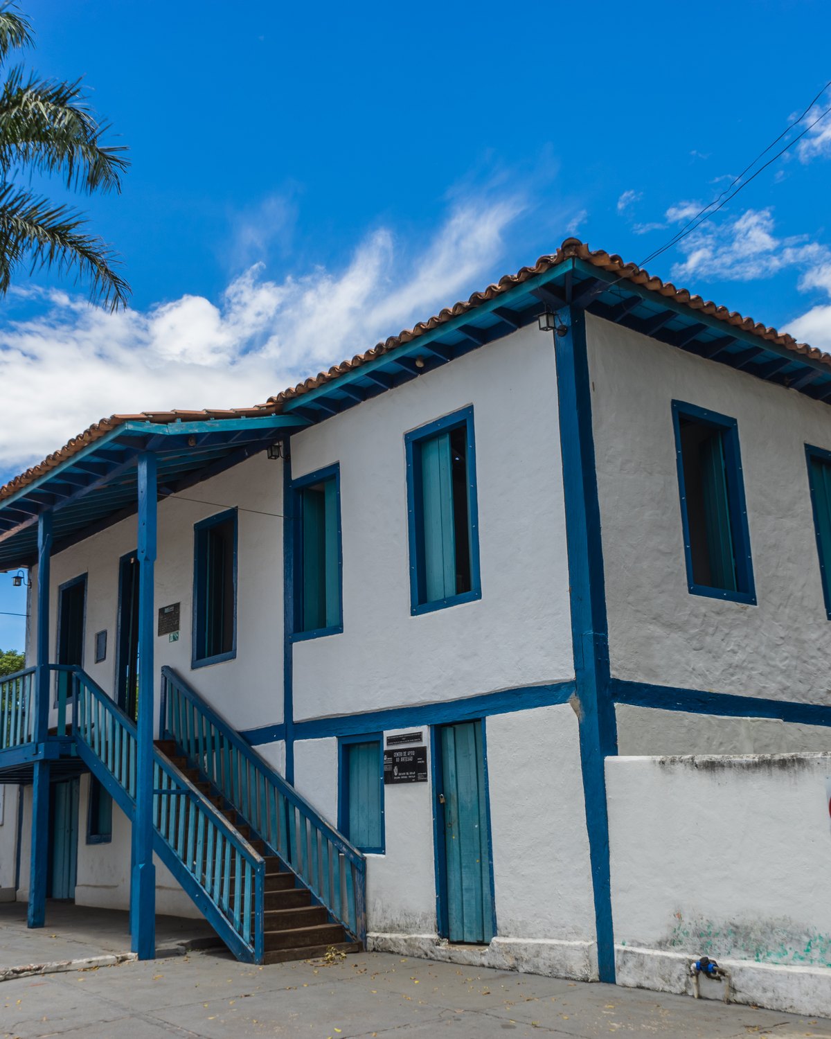 Casarão dos Gonzaga, sobrado branco de janelas e varanda azuis com escada lateral de madeira, numa esquina do Centro de Caldas Novas, com palmeiras e céu azul.