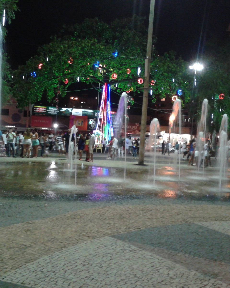Praça no Centro de Caldas Novas à noite, com jatos de água saindo do piso de pedra portuguesa, árvores enfeitadas com luzes coloridas de fim de ano e muita gente passeando ao fundo.