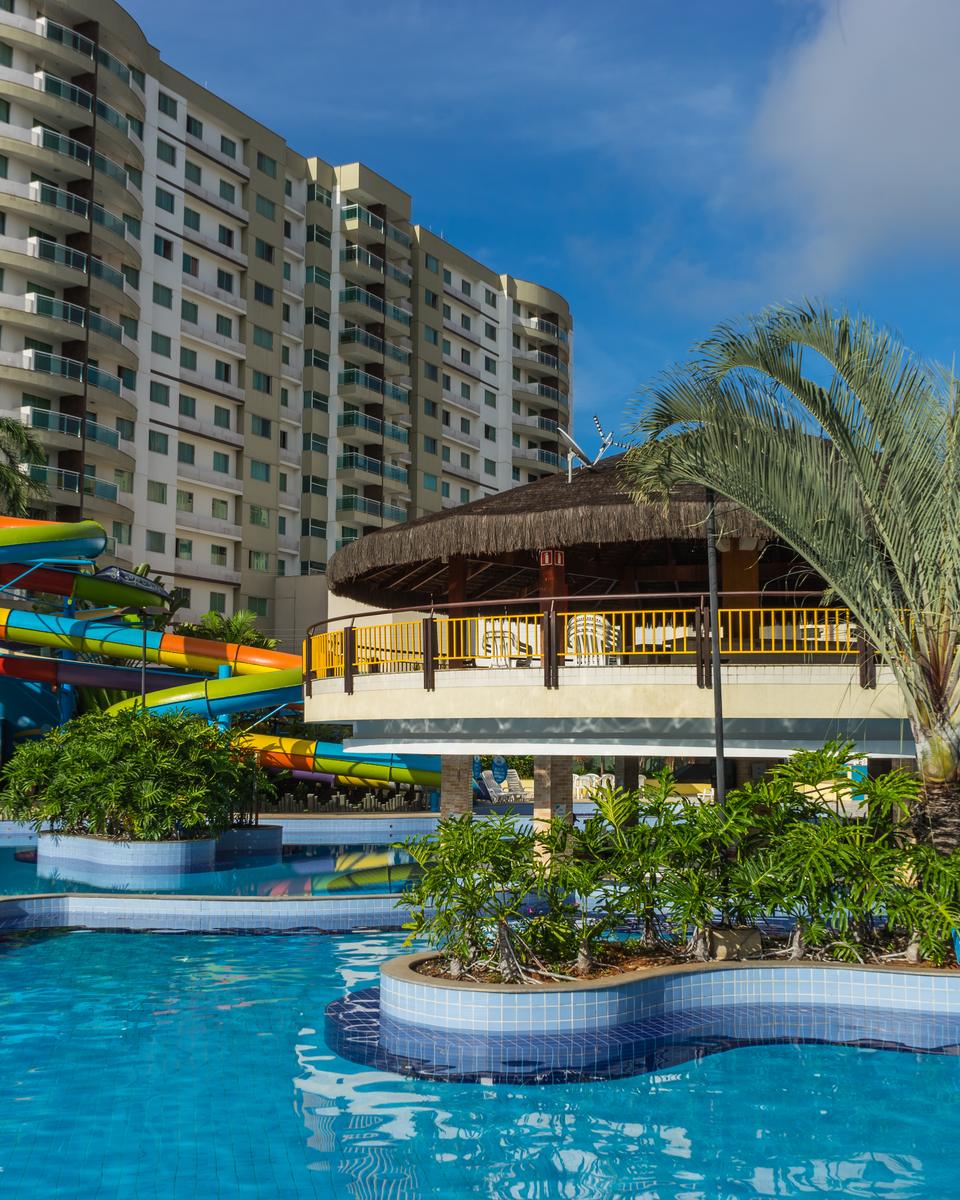 Área de lazer de um clube em Caldas Novas, com piscina azul, ilhas de palmeiras, quiosque de palha, toboáguas coloridos e torres altas de apartamentos de temporada ao fundo, sob céu azul.