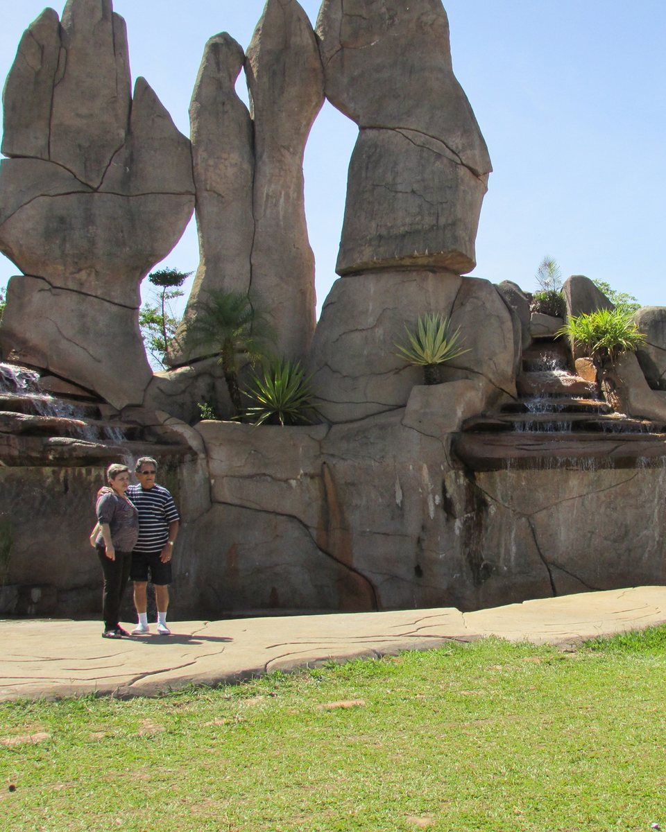 Casal de turistas posando para foto diante das grandes esculturas de pedra e da cascata do Monumento das Águas, em Caldas Novas, num dia de céu azul.