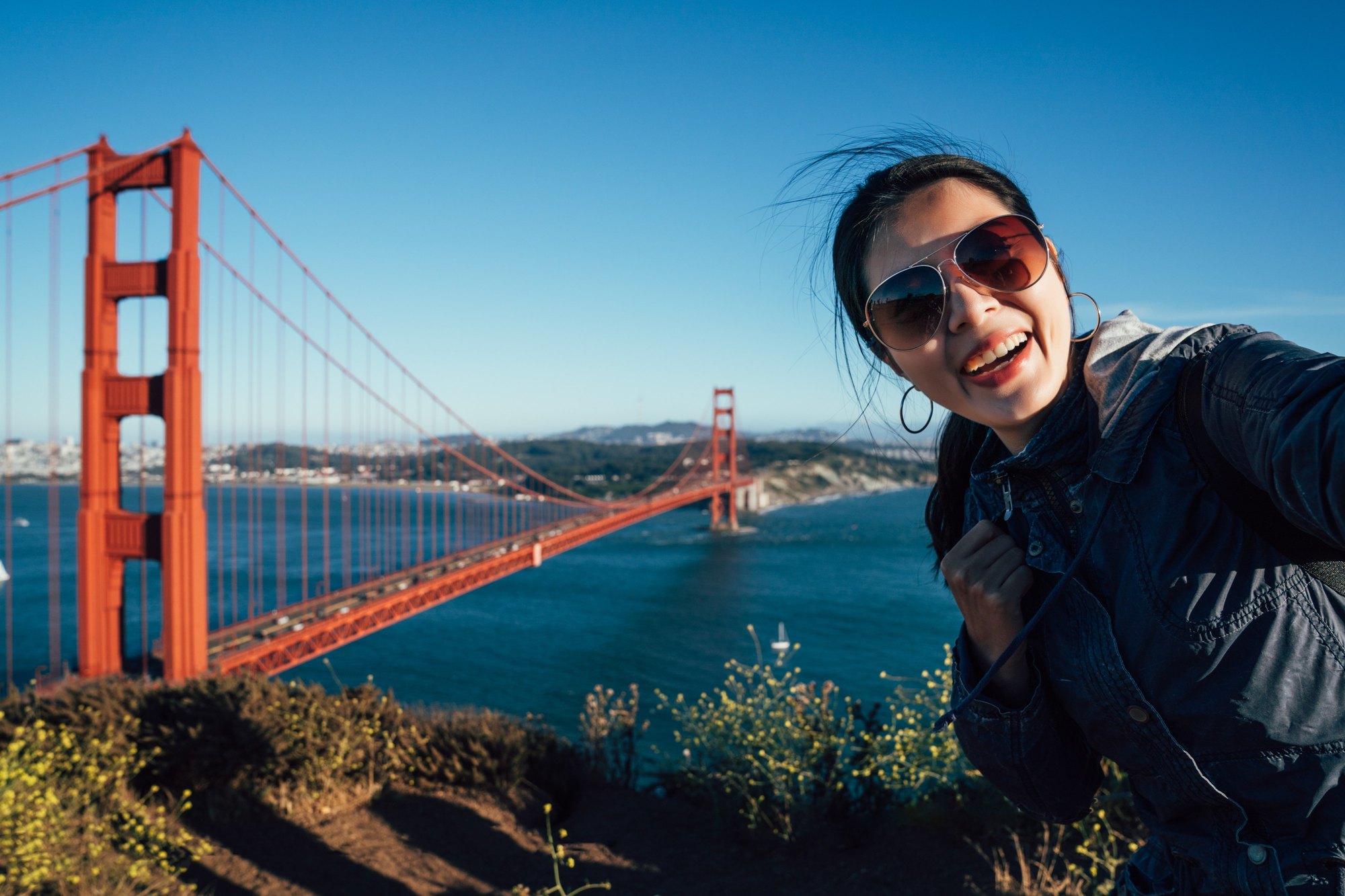 Jovem de óculos escuros sorrindo e tirando selfie com o celular num mirante, com a Golden Gate Bridge e a baía de San Francisco ao fundo.