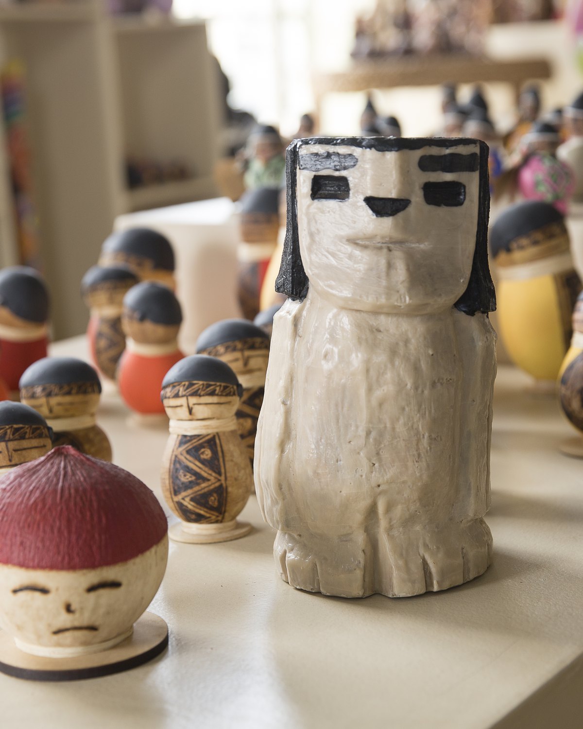 Bugre esculpido à mão em destaque sobre uma bancada branca da Casa do Artesão, cercado por pequenas peças arredondadas de artesanato coloridas.